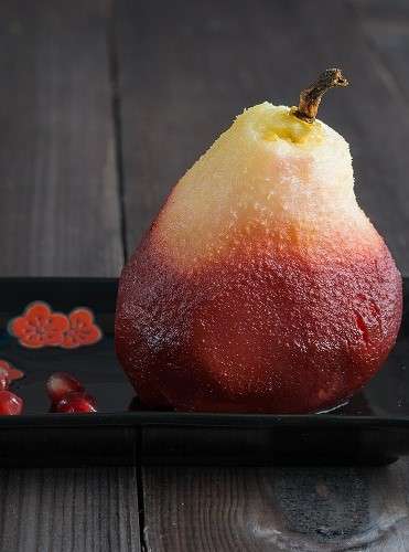 one poached pear on black rectangle plate with red pomegranate seeds on dark wooden background